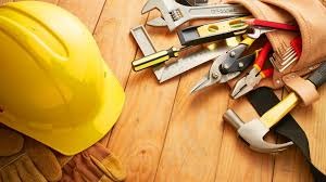 Żółty kask ochronny, rękawice robocze i pas z narzędziami (klucze, poziomica, nożyce do blachy, młotek) ułożone na drewnianym blacie.