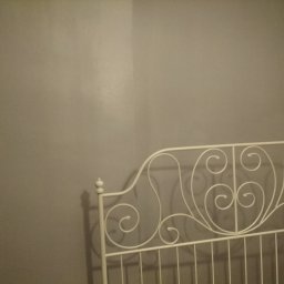 Sabinex - Fragment metalowego, białego wezgłowia łóżka z dekoracyjnymi zawijasami na tle szarej ściany w słabym oświetleniu.