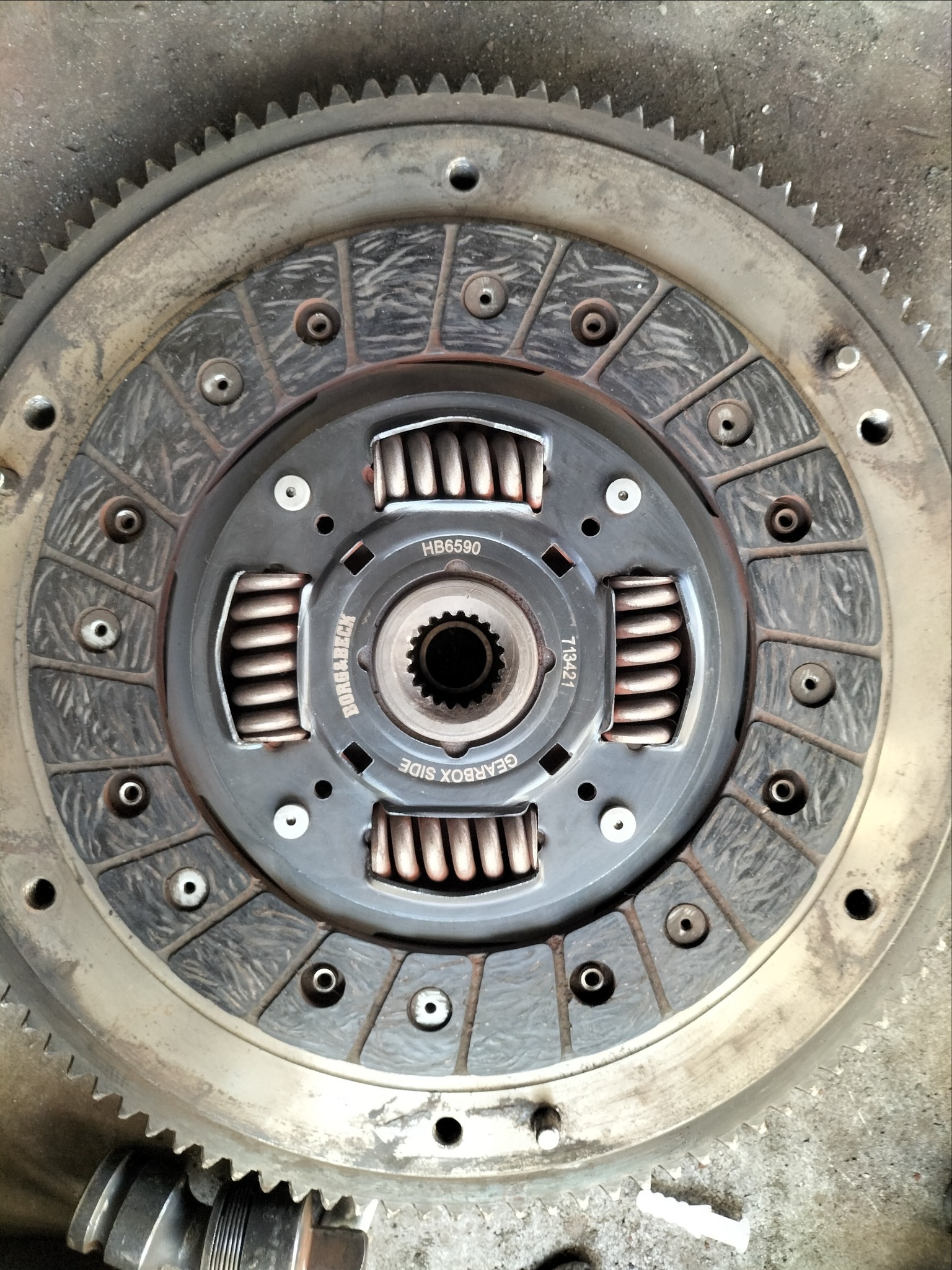 Tarcza sprzęgła Borg & Beck HB6590 z widocznymi sprężynami i oznaczeniem 'GEARBOX SIDE', osadzona w kole zamachowym z zębami, na brudnym tle.
