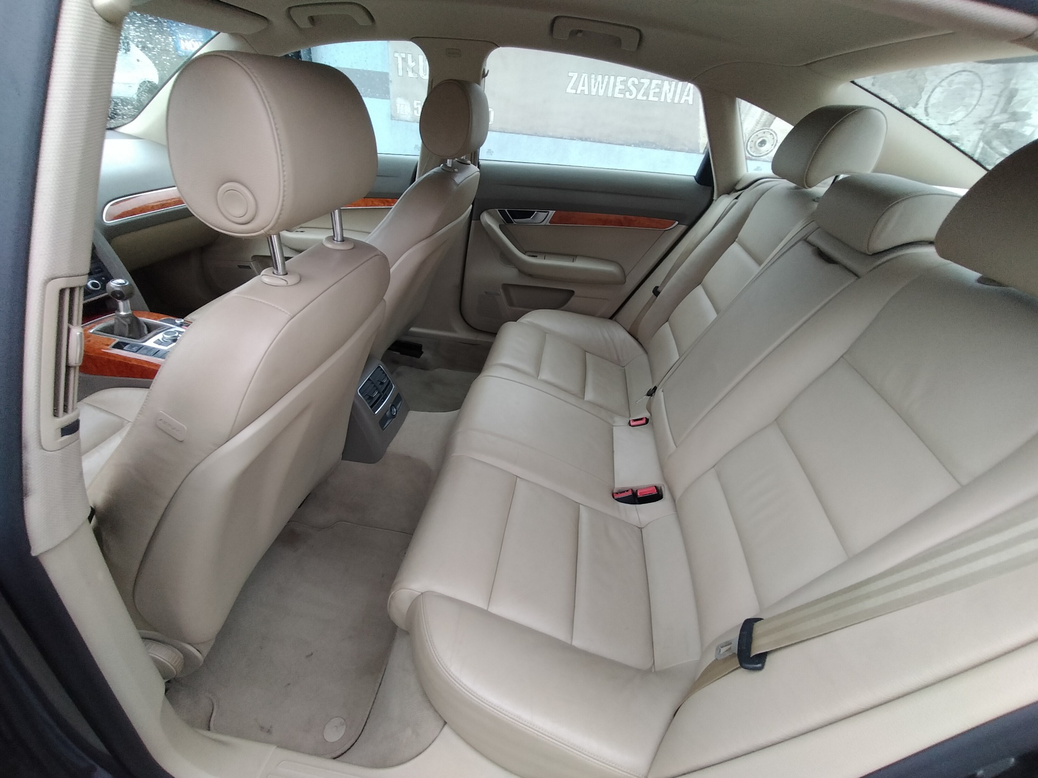 Wnętrze samochodu z beżową skórzaną tapicerką, widok na tylne siedzenia z zamontowanymi pasami bezpieczeństwa oraz fragment deski rozdzielczej z elementami drewnopodobnymi.