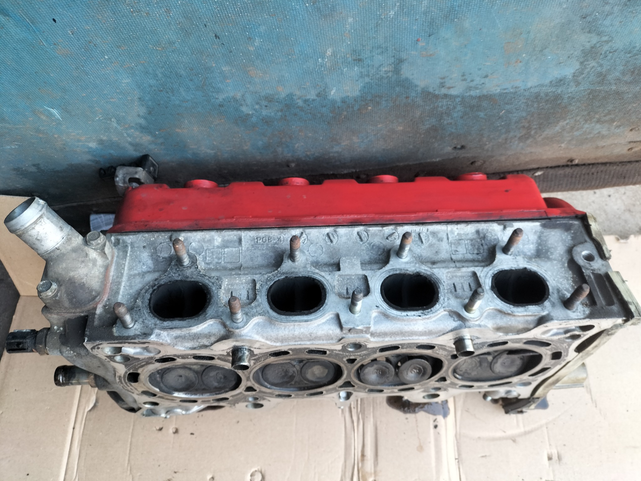 Głowica silnika spalinowego, widoczne zawory, kanały i czerwona pokrywa, leżąca na kartonie.