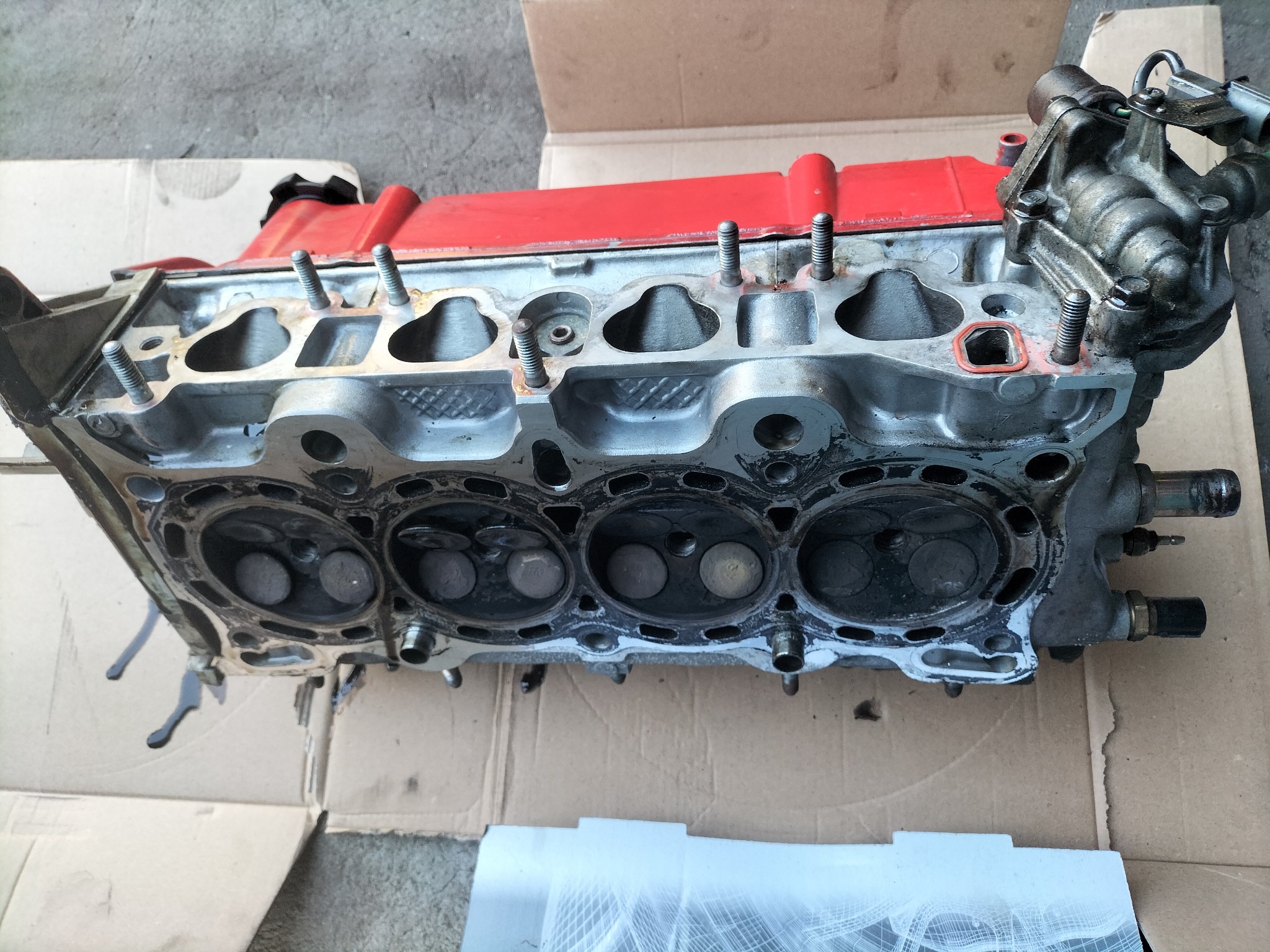 Zdemontowana głowica silnika samochodowego z czerwonym deklem, widoczne zawory i ślady oleju, leżąca na kartonie.