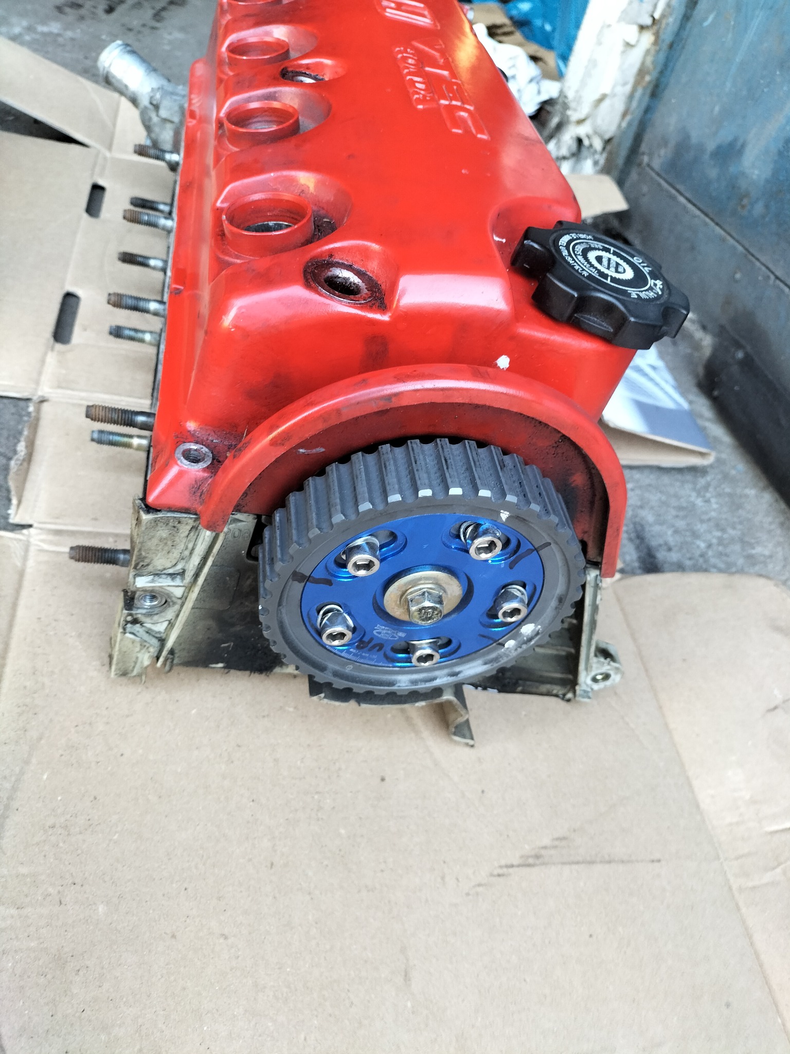 Czerwona głowica silnika z niebieskim kołem zębatym na tekturowym podłożu, widoczne ślady użytkowania i zabrudzenia.