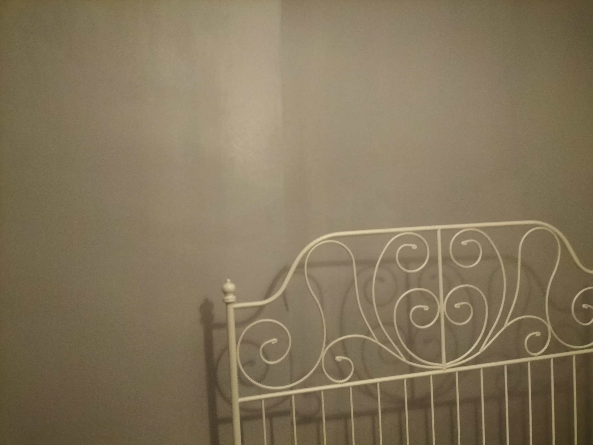 Fragment metalowego, białego wezgłowia łóżka z dekoracyjnymi zawijasami na tle szarej ściany w słabym oświetleniu.
