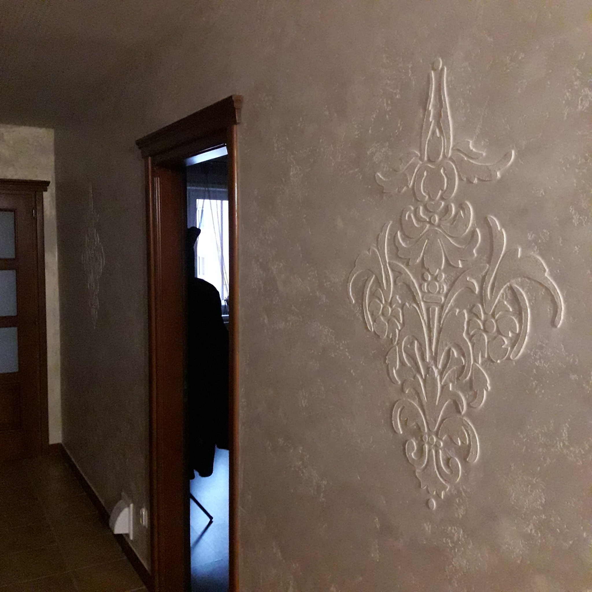 Dekoracyjna ściana w beżowym odcieniu z wypukłym, symetrycznym ornamentem w stylu rokoko, widok z perspektywy wnętrza mieszkania, z widoczną ościeżnicą drzwi i fragmentem innego pomieszczenia.