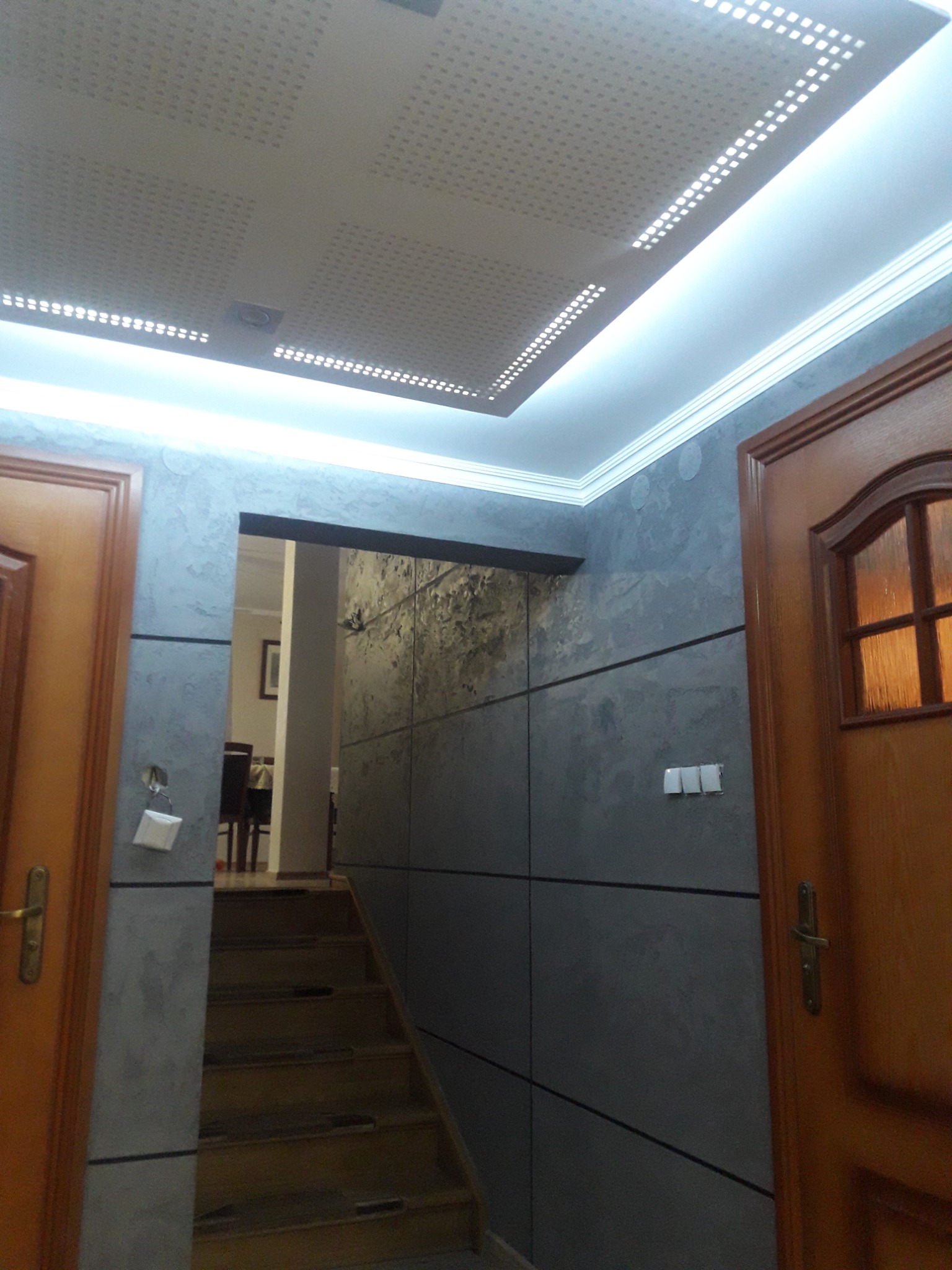 Korytarz z szarą, strukturalną ścianą, drewnianymi drzwiami po bokach i schodami prowadzącymi w górę, oświetlony listwami LED pod sufitem z perforowanymi panelami.