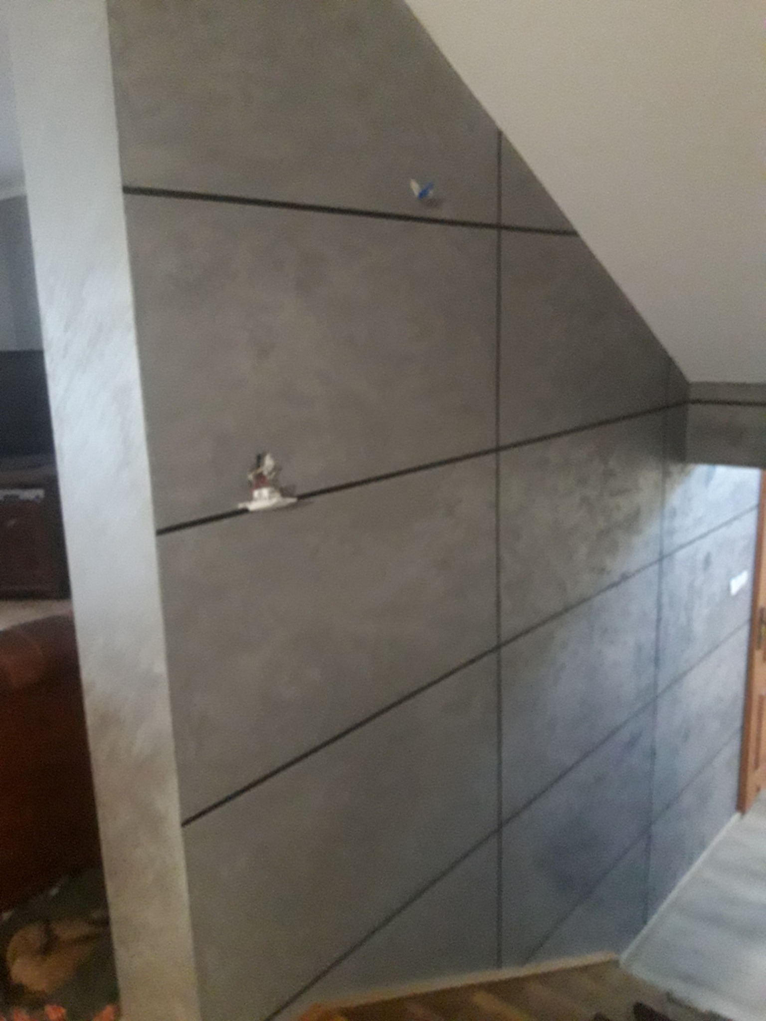 Ściana w szarym odcieniu z czarnymi liniami tworzącymi geometryczny wzór, widoczne elementy instalacji elektrycznej, fragment podłogi i drzwi w tle.