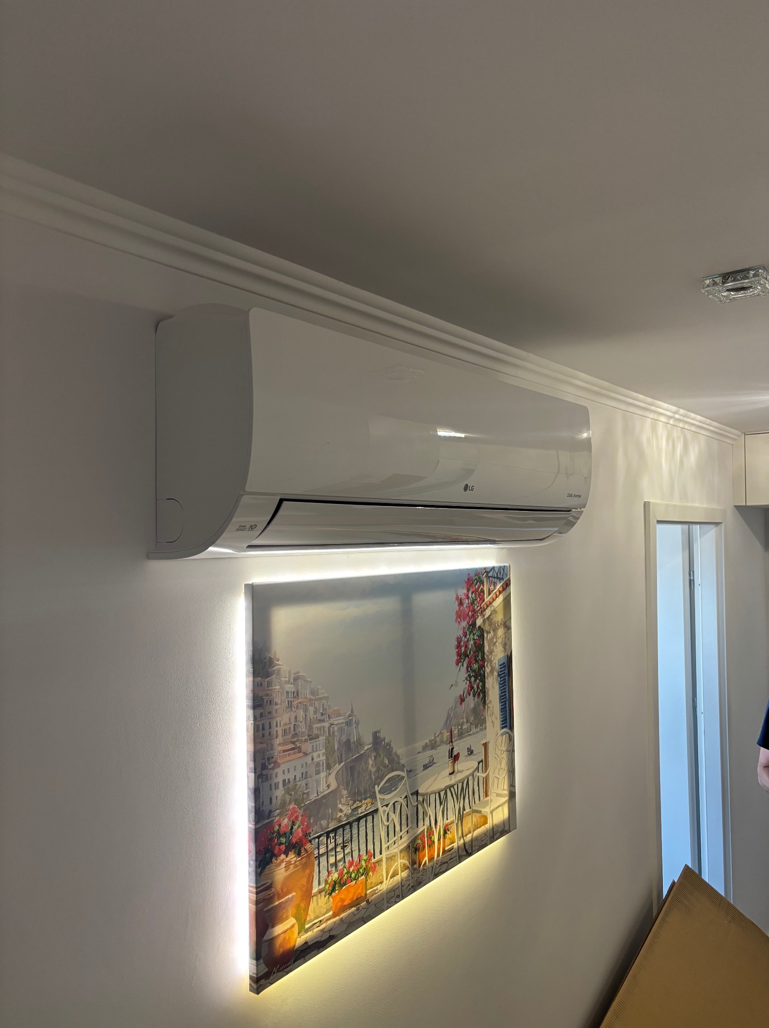 Biała klimatyzacja LG na ścianie nad podświetlanym obrazem z widokiem na włoskie miasteczko. Nowoczesny design i oświetlenie tworzą przyjemny klimat we wnętrzu.