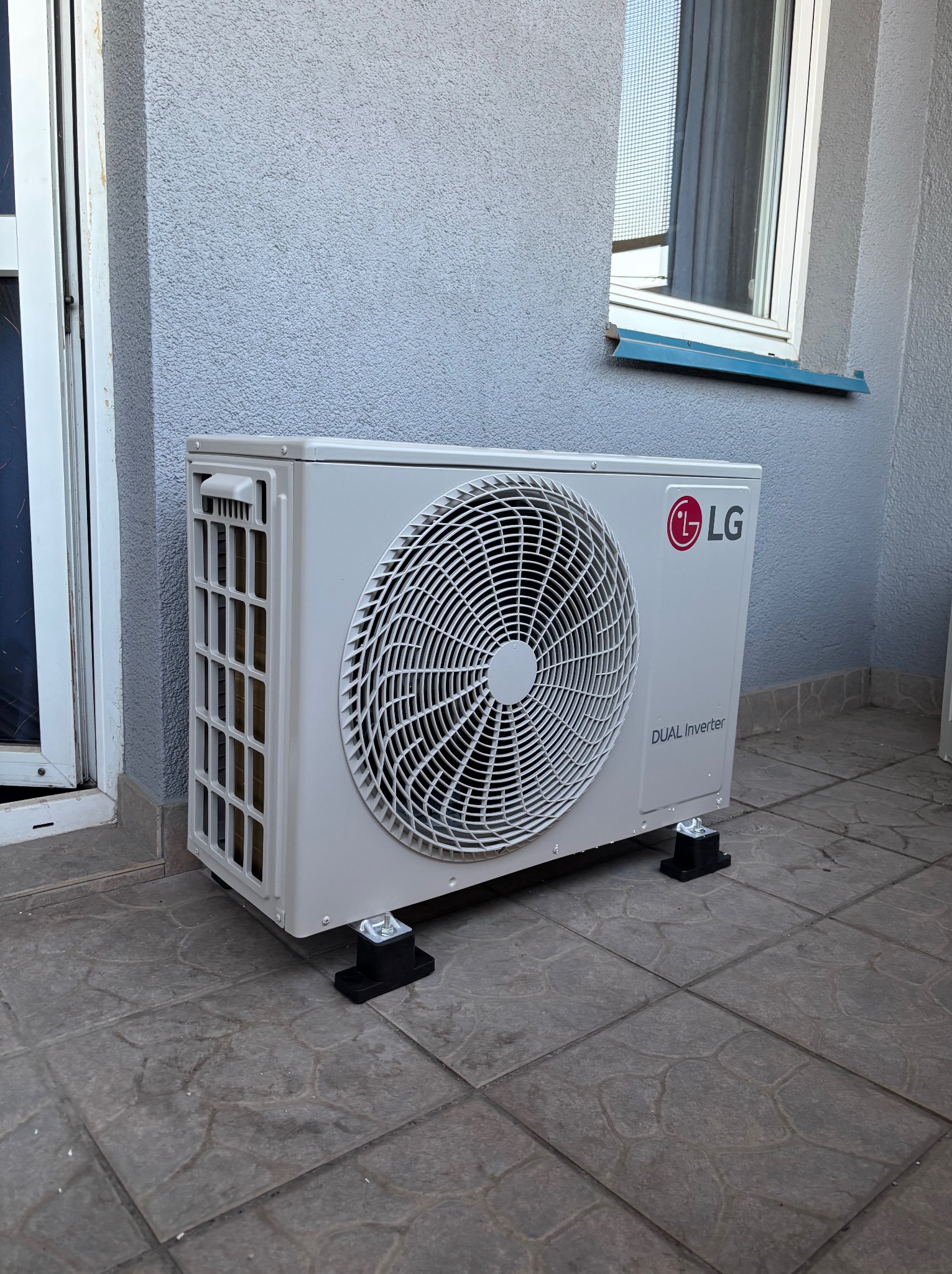 Zewnętrzna jednostka klimatyzacji LG Dual Inverter, zamontowana na czarnych podstawkach na szarej, wzorzystej posadzce, przy szarej ścianie z oknem i drzwiami.