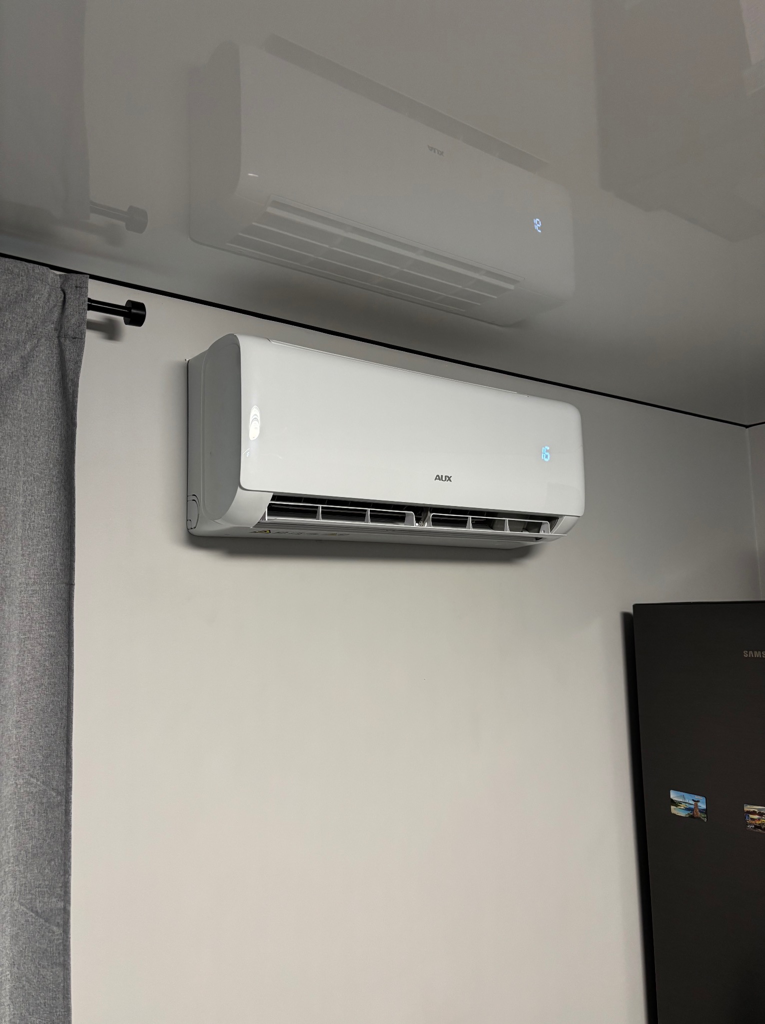 Biała klimatyzacja AUX na ścianie w pomieszczeniu, widoczny wyświetlacz z temperaturą 16 stopni, zasłona i fragment lodówki. Widoczne odbicie klimatyzacji w suficie.