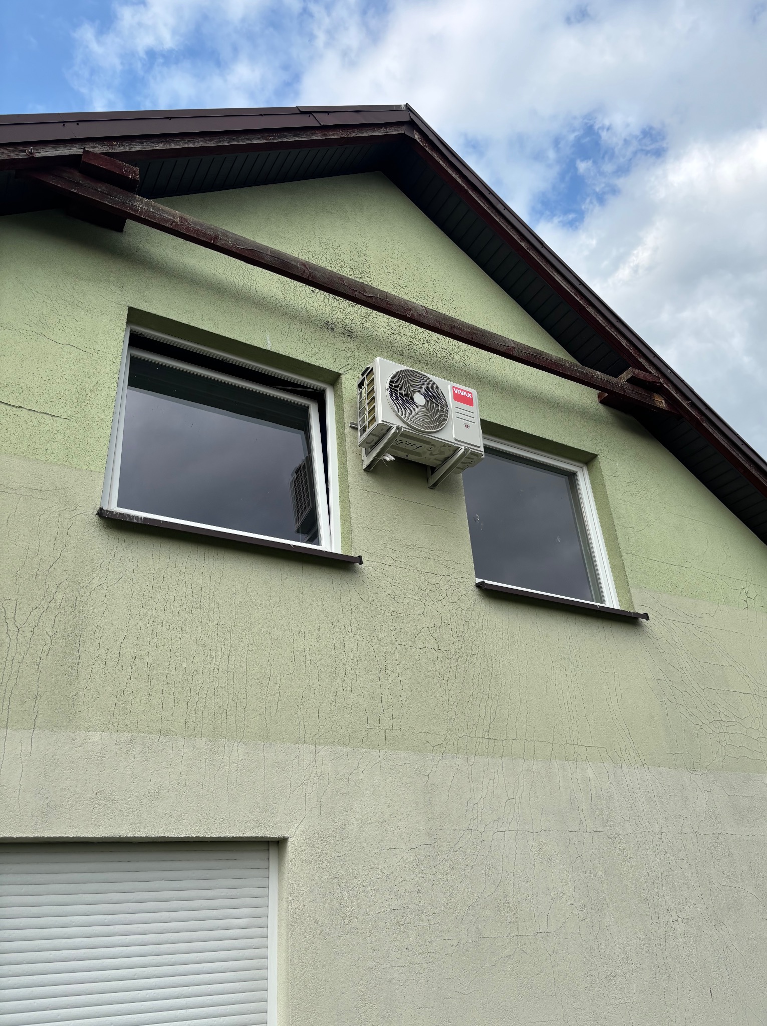 Zewnętrzna jednostka klimatyzacji VIVAX zamontowana na elewacji domu o zielonym odcieniu, widoczna między dwoma oknami. Tło stanowi błękitne niebo z chmurami.