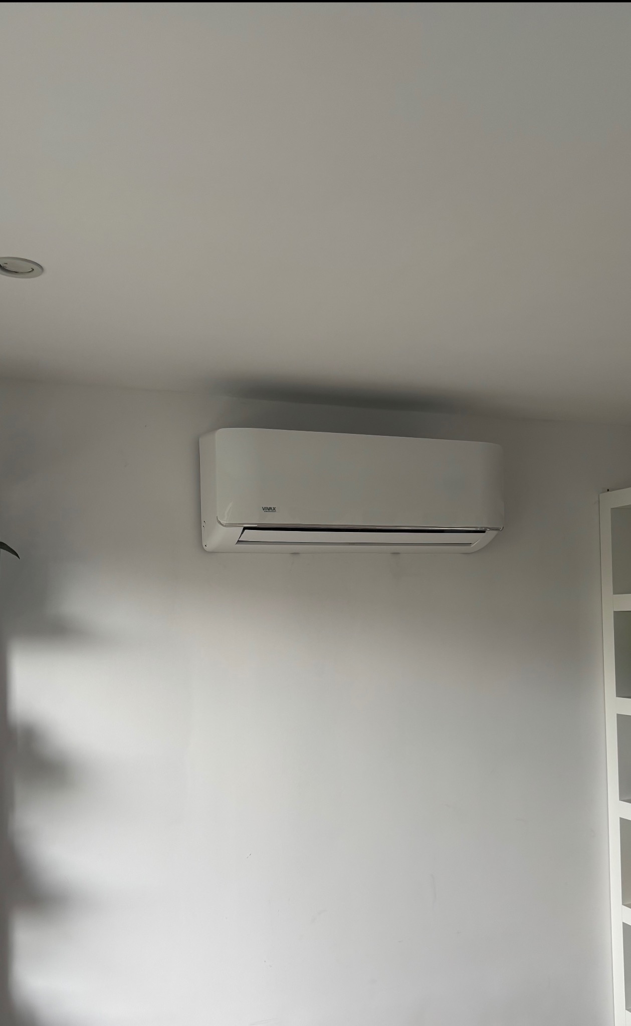 Biała klimatyzacja VIVAX zamontowana na jasnej ścianie w pomieszczeniu. Widoczny fragment regału po prawej stronie. Minimalistyczny design wnętrza.
