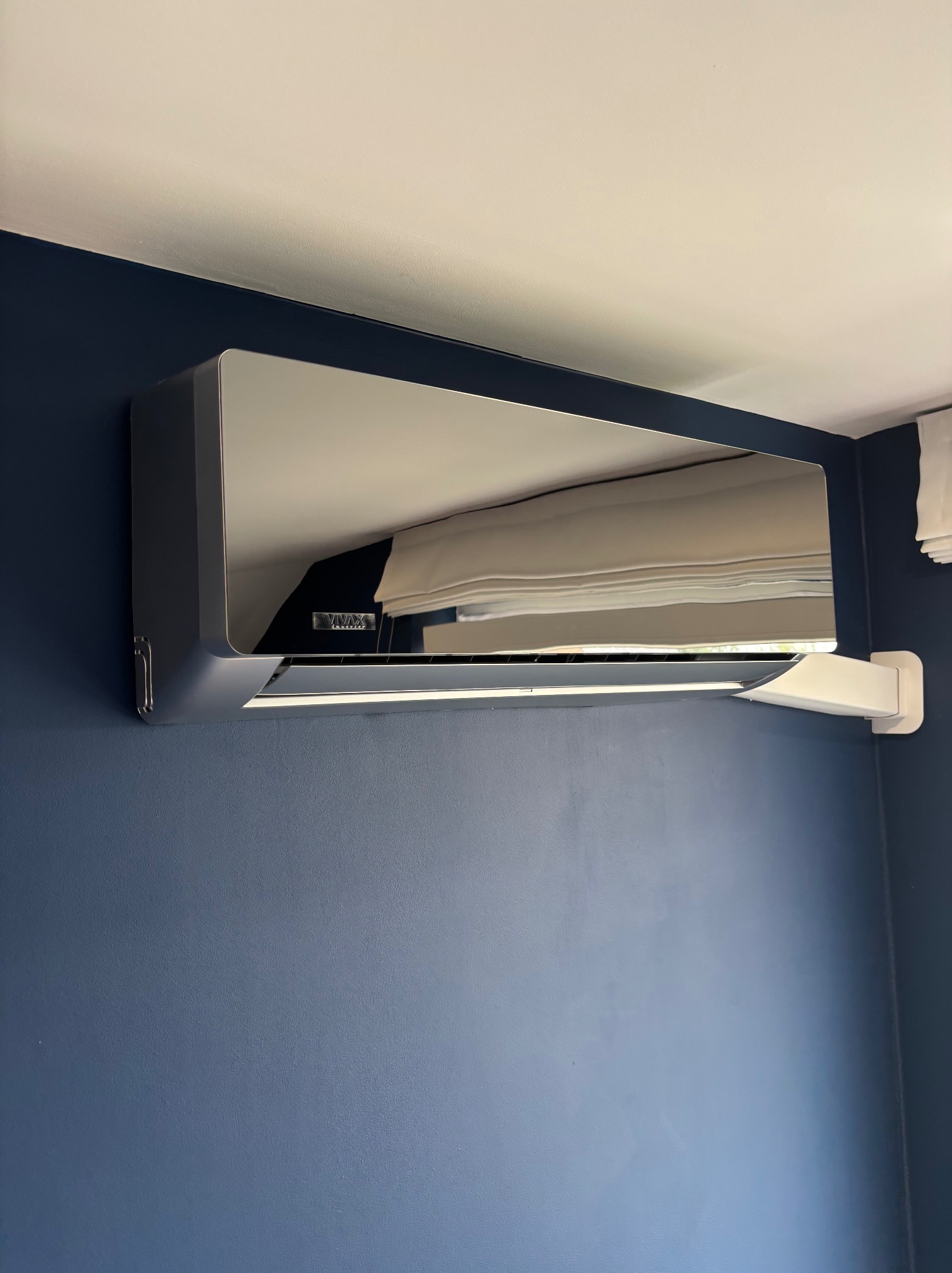 Błyszczący, srebrny klimatyzator VIVAX zamontowany na granatowej ścianie, z widocznym odbiciem wnętrza. Nowoczesny design, eleganckie wykończenie.