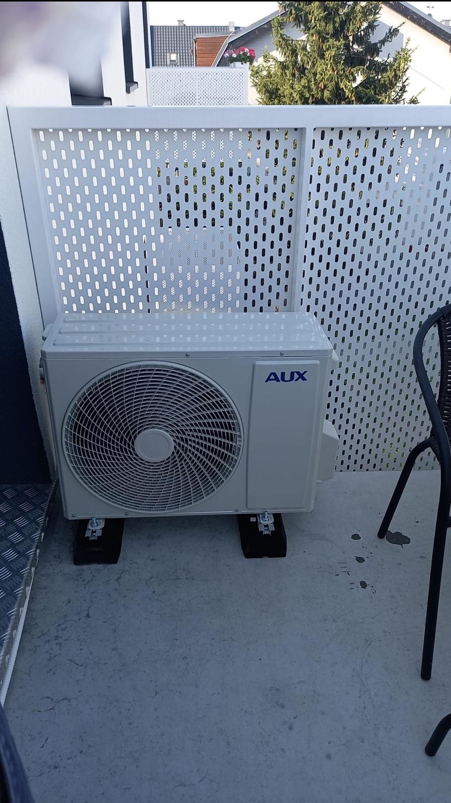 Biały klimatyzator AUX na balkonie, ustawiony na czarnych podkładkach, z ażurową balustradą w tle i fragmentem krzesła. Widok z zewnątrz.