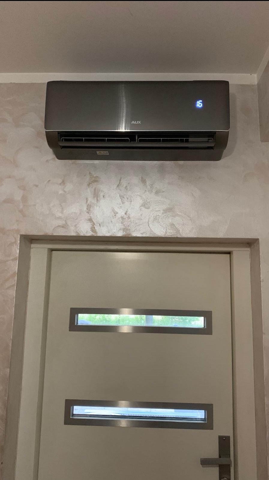 Szara klimatyzacja AUX zamontowana nad drzwiami we Wrocławiu. Na ścianie dekoracyjny tynk. Widoczny wyświetlacz temperatury na urządzeniu.