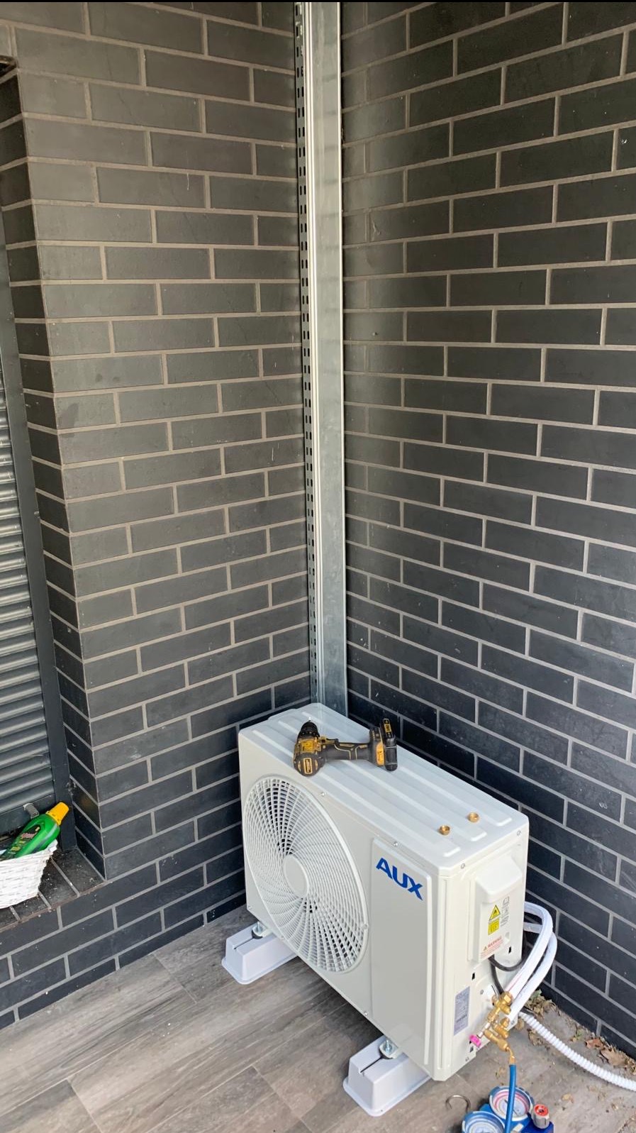Biała jednostka zewnętrzna klimatyzacji AUX na szarym podłożu, obok ciemnej ściany z cegły. Na urządzeniu wiertarko-wkrętarka. Widoczne przewody i manometry.