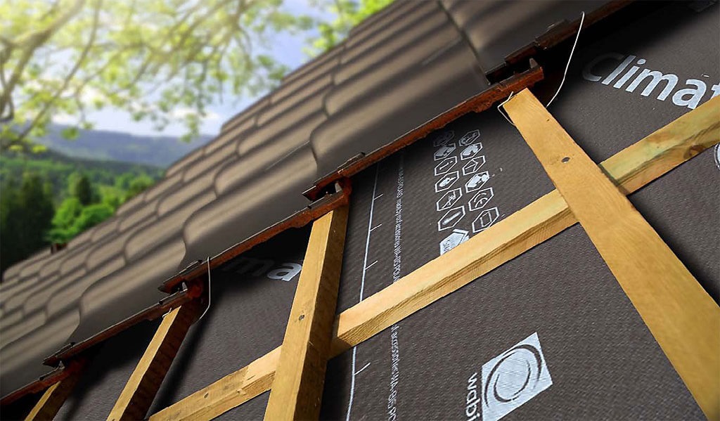 Fragment dachu z brązową dachówką ceramiczną, widoczne drewniane łaty i membrana dachowa z nadrukiem, w tle zieleń drzew i wzgórz.