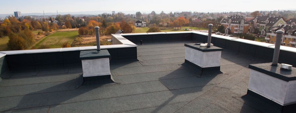 Widok z góry na płaski dach pokryty papą termozgrzewalną z kominami wentylacyjnymi, na tle zabudowy miejskiej i jesiennego krajobrazu.