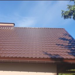 Uslugi dekarskie - Brązowy dach z blachodachówki trapezowej, widoczne rynny w tym samym kolorze, komin i fragment ściany budynku na tle błękitnego nieba i gałęzi drzewa iglastego.