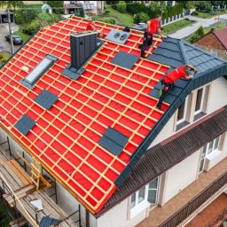 Uslugi dekarskie - Ekipa dekarzy w czerwonych uniformach montuje ciemnoszarą dachówkę na nowym dachu domu jednorodzinnego z widoczną czerwoną membraną, oknem dachowym i kominem.