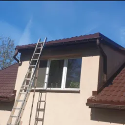 Dom jednorodzinny z brązowym dachem z blachodachówki, brązowymi rynnami i drabiną opartą o ścianę przy oknie, na tle błękitnego nieba i zieleni drzew.