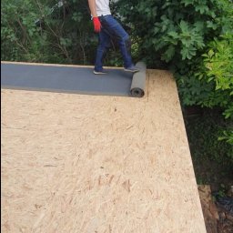 Uslugi dekarskie - Pracownik w czerwonych rękawicach rozkłada rolkę czarnej papy na drewnianym poszyciu dachu, w tle zielona roślinność.