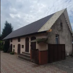 Częściowo wymieniony dach na starym budynku gospodarczym, widoczne nowe brązowe dachówki i membrana dachowa na szczycie dachu, ceglana ścieżka prowadząca do wejścia.