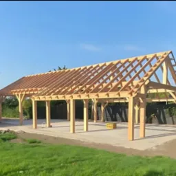 Wznoszenie konstrukcji dachu z drewnianych belek na betonowej wylewce, widoczne elementy łączące i zielone otoczenie.