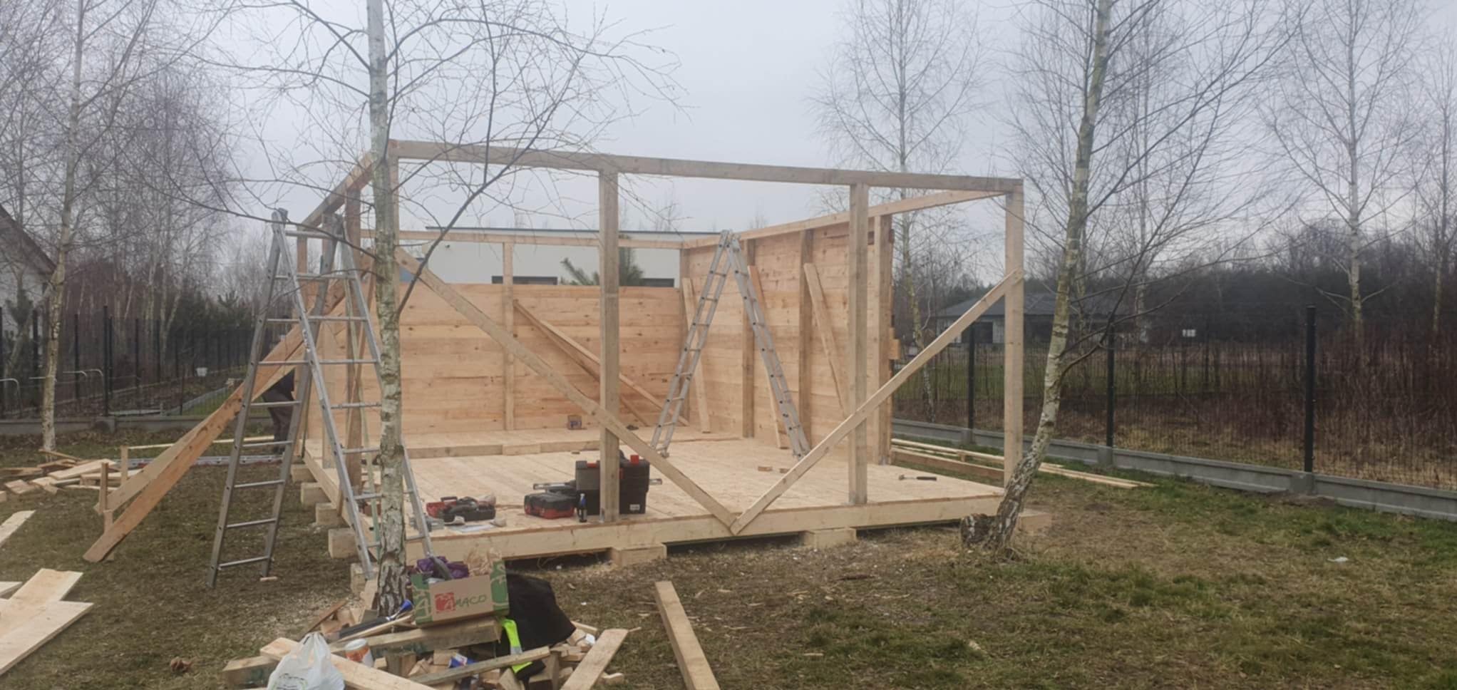 Konstrukcja szkieletowa drewnianej altany w trakcie budowy, widoczne narzędzia, drabiny i materiały budowlane na trawie, pochmurne niebo.