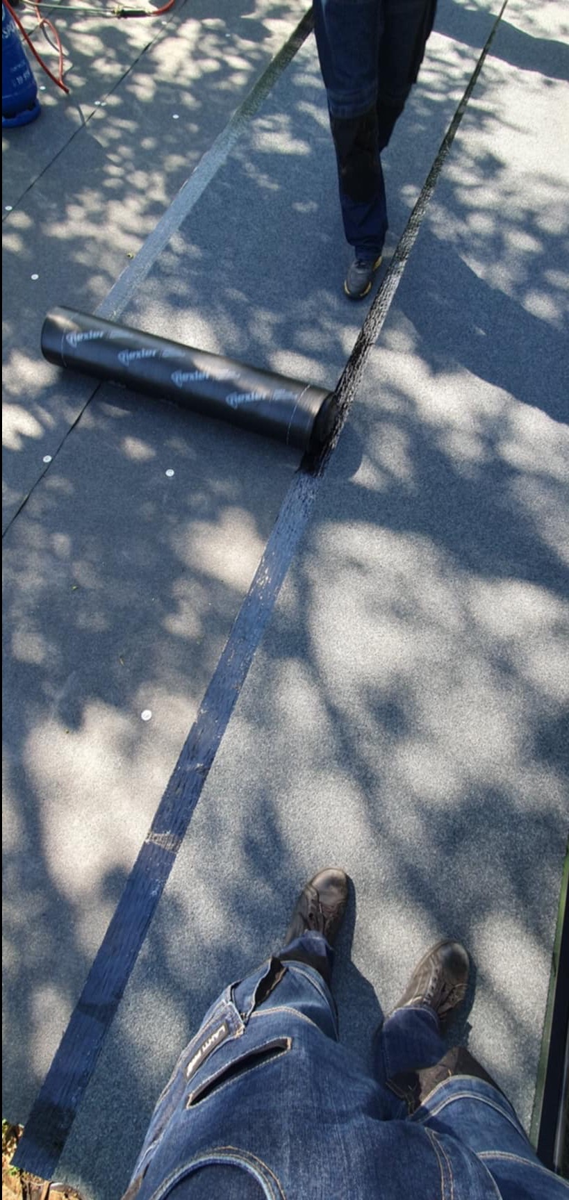 Widok z góry na fragment dachu z położoną papą termozgrzewalną, częściowo rozwiniętą z rolki marki Bester, z widocznymi cieniami drzew i stopami dekarza w jeansach i roboczych butach.