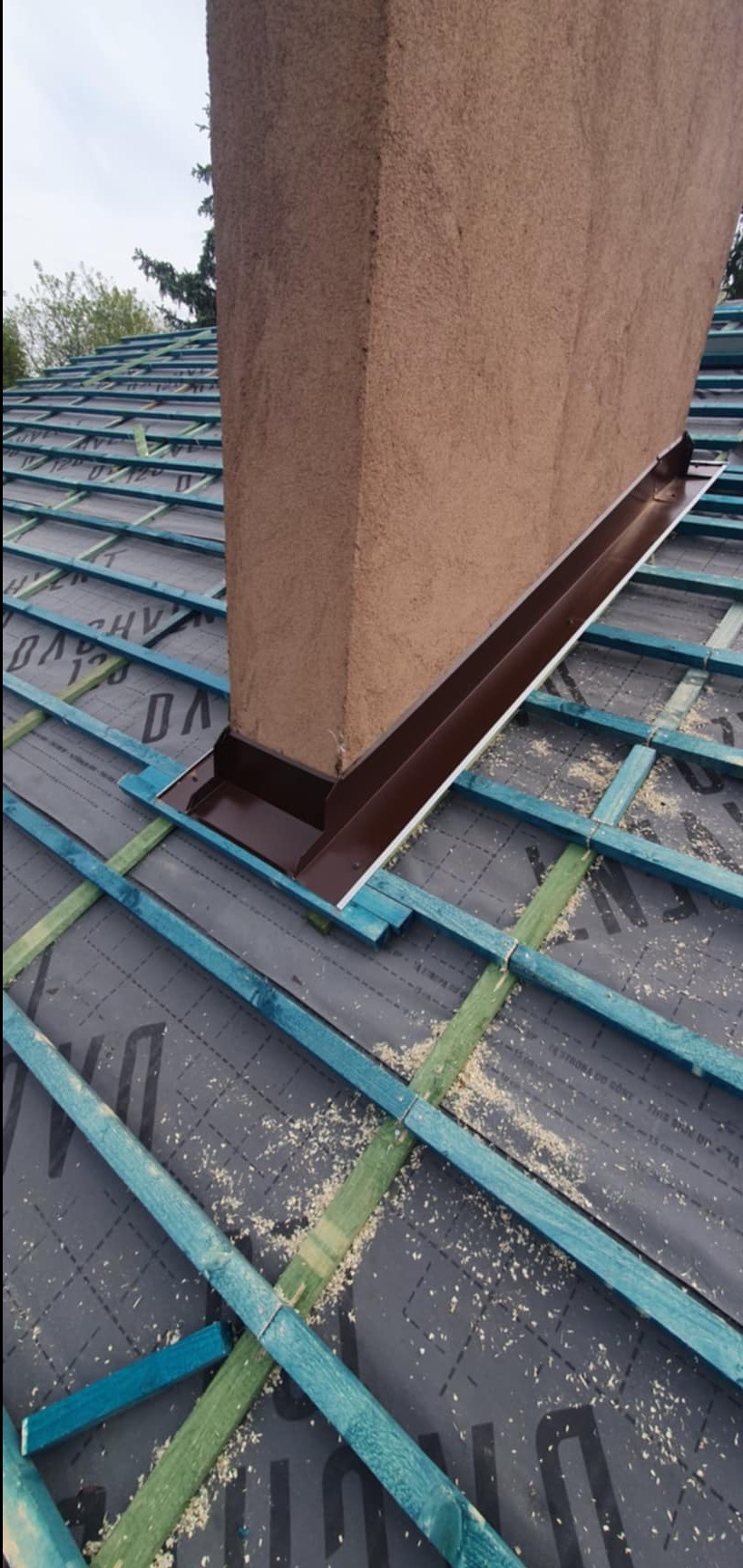 Fragment dachu w trakcie budowy z kominem zabezpieczonym brązową obróbką blacharską, widoczna impregnacja więźby dachowej i membrana dachowa.