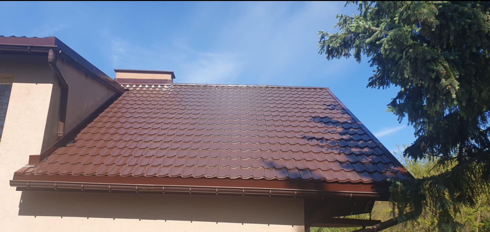 Brązowy dach z blachodachówki trapezowej, widoczne rynny w tym samym kolorze, komin i fragment ściany budynku na tle błękitnego nieba i gałęzi drzewa iglastego.