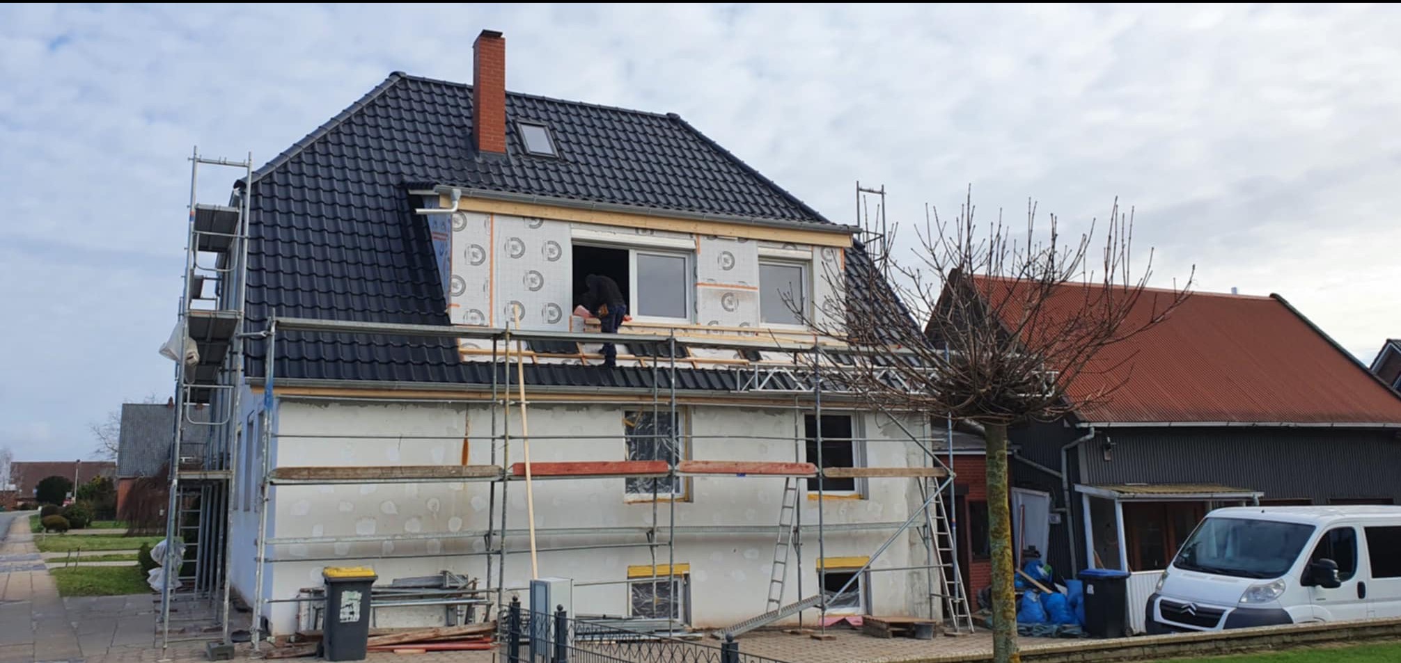 Dom jednorodzinny w trakcie ocieplania poddasza i wymiany pokrycia dachowego, widoczne rusztowania, nowy czarny dach z oknem dachowym i kominem, oraz pracownik w oknie poddasza.