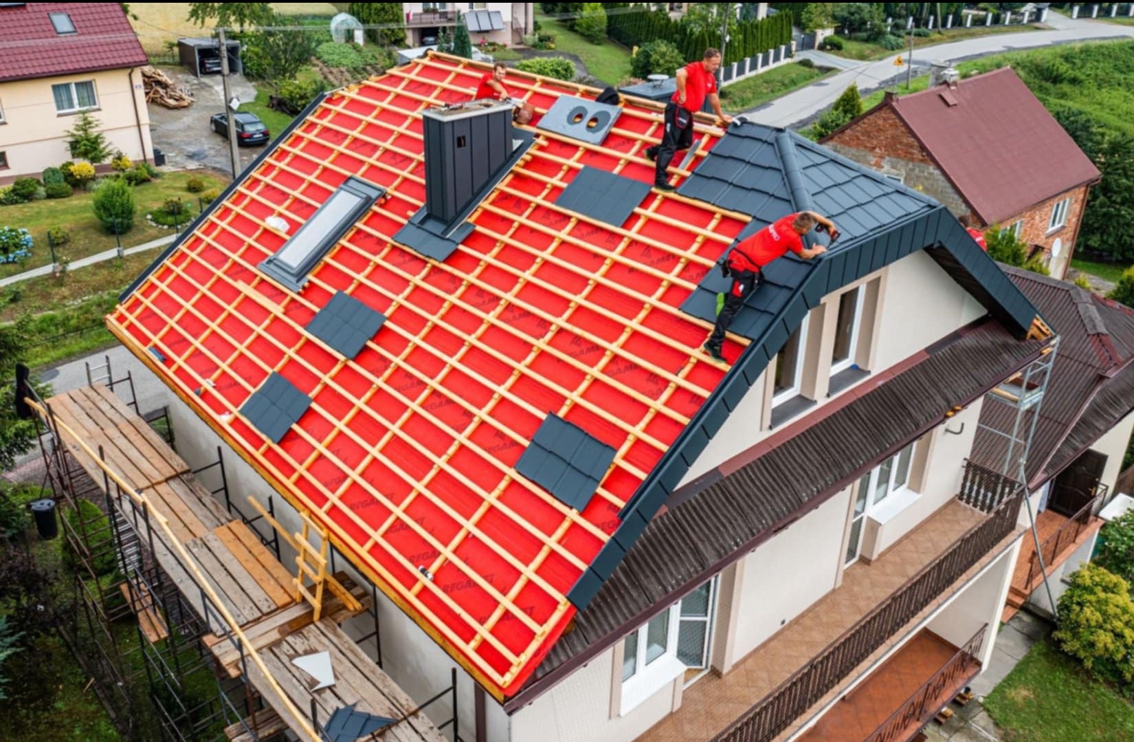 Ekipa dekarzy w czerwonych uniformach montuje ciemnoszarą dachówkę na nowym dachu domu jednorodzinnego z widoczną czerwoną membraną, oknem dachowym i kominem.
