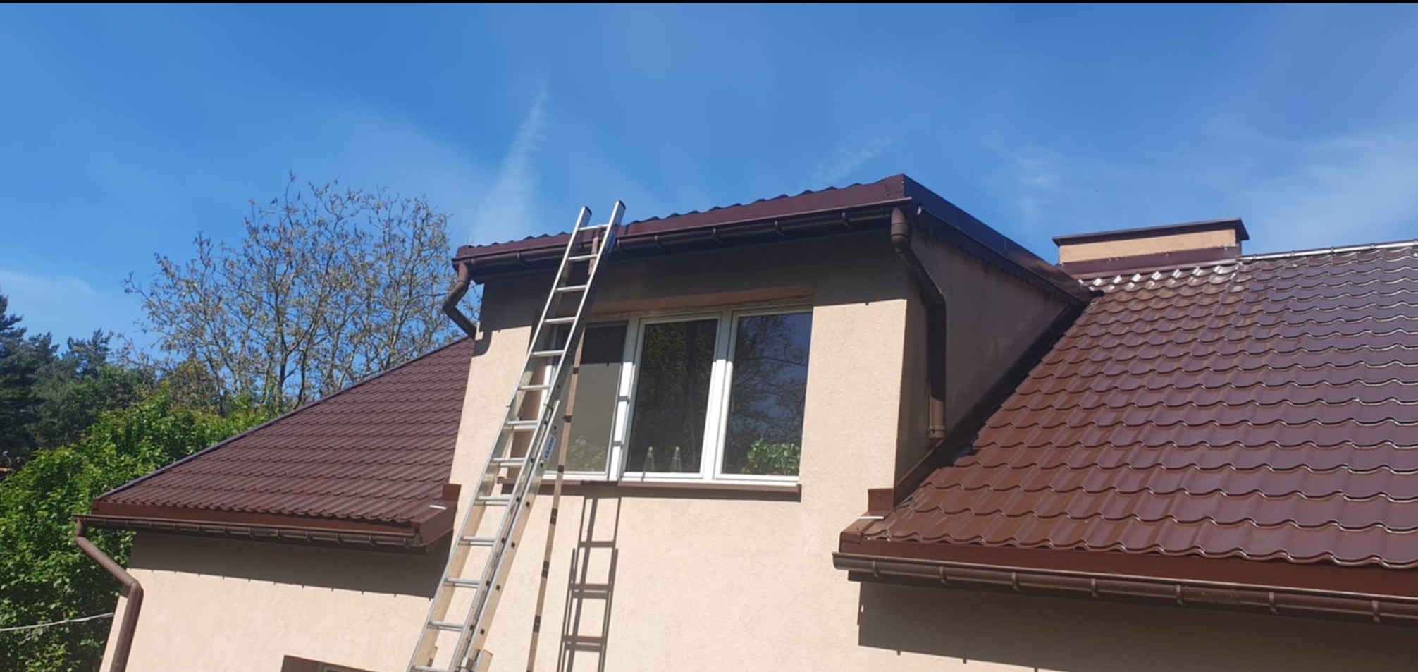 Dom jednorodzinny z brązowym dachem z blachodachówki, brązowymi rynnami i drabiną opartą o ścianę przy oknie, na tle błękitnego nieba i zieleni drzew.