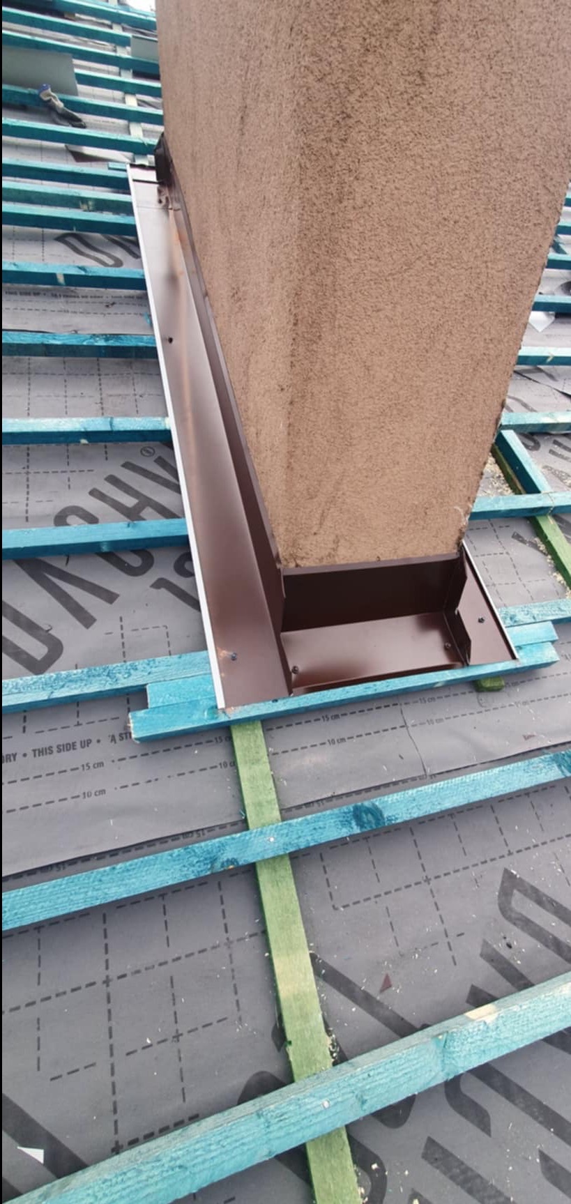 Szczegółowe ujęcie brązowej obróbki blacharskiej komina na dachu pokrytym czarną membraną z widoczną drewnianą konstrukcją więźby pomalowaną na niebiesko-zielony kolor.