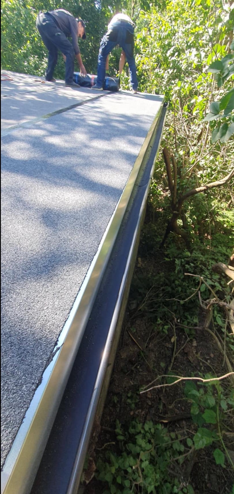 Ekipa dekarzy układa papę termozgrzewalną na dachu z zamontowaną, metalową rynną w otoczeniu zieleni.