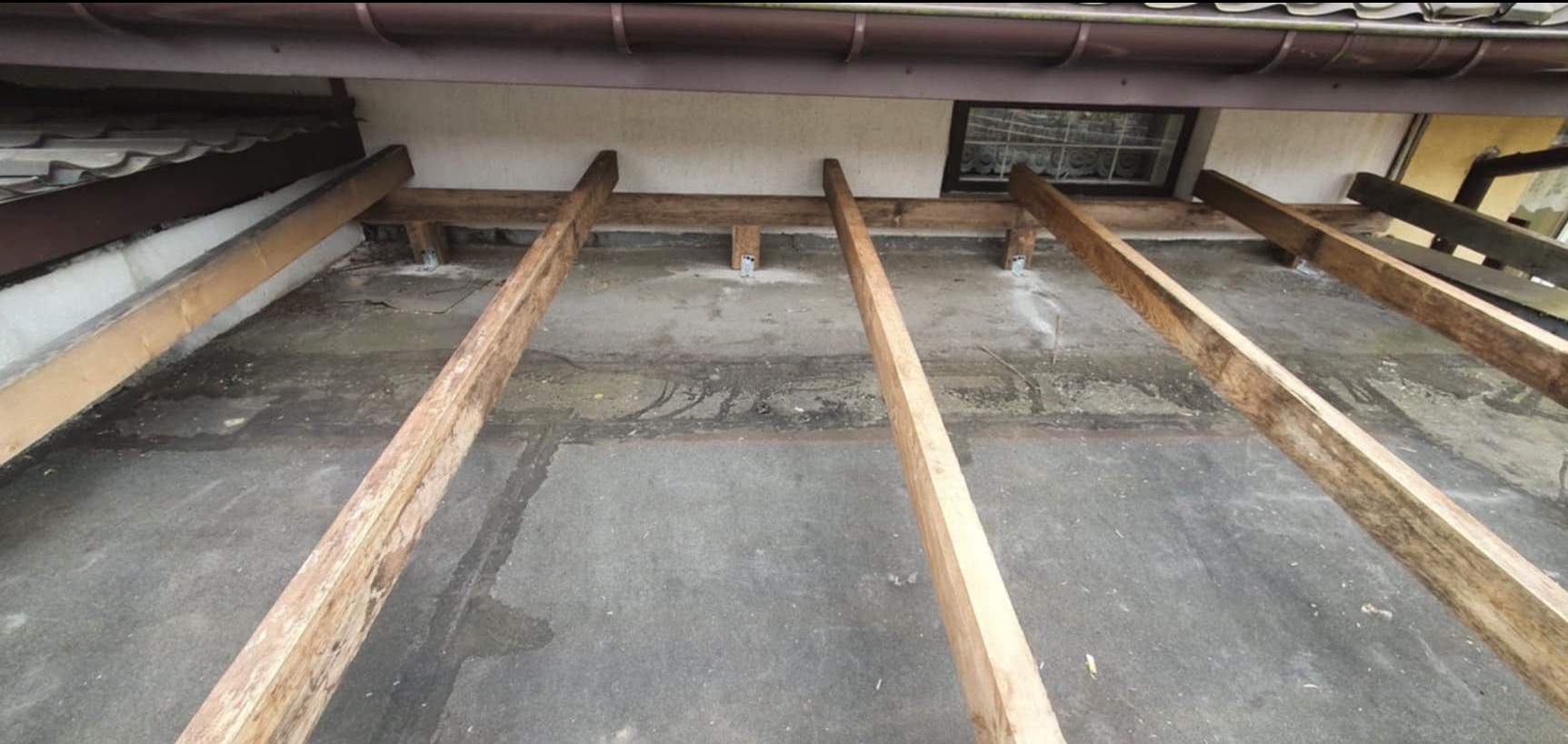 Drewniana konstrukcja więźby dachowej z metalowymi mocowaniami, widoczna spod spodu, z fragmentem dachu i rynny w tle.