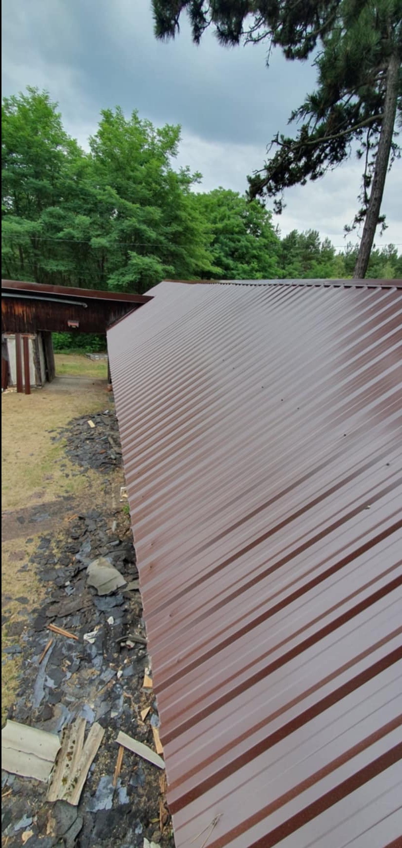 Nowy, brązowy dach z blachy trapezowej na budynku gospodarczym, widok z góry na tle zieleni drzew i pochmurnego nieba. Obok dachu widoczne pozostałości starego pokrycia.