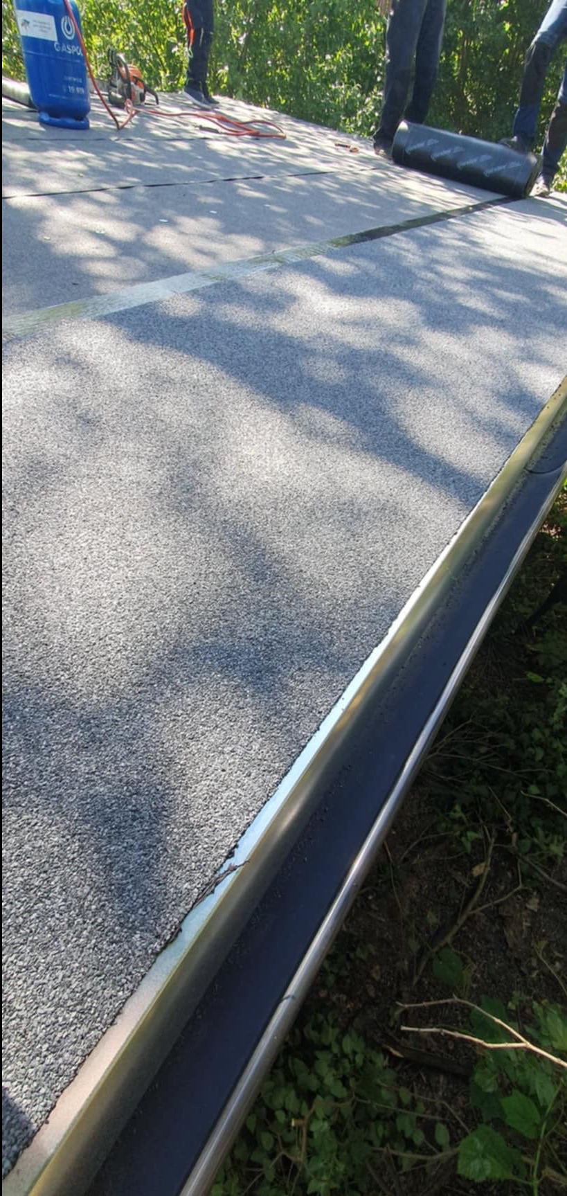 Fragment dachu pokrytego papą termozgrzewalną, z widoczną nową rynną metalową, butla gazowa i narzędzia w tle.