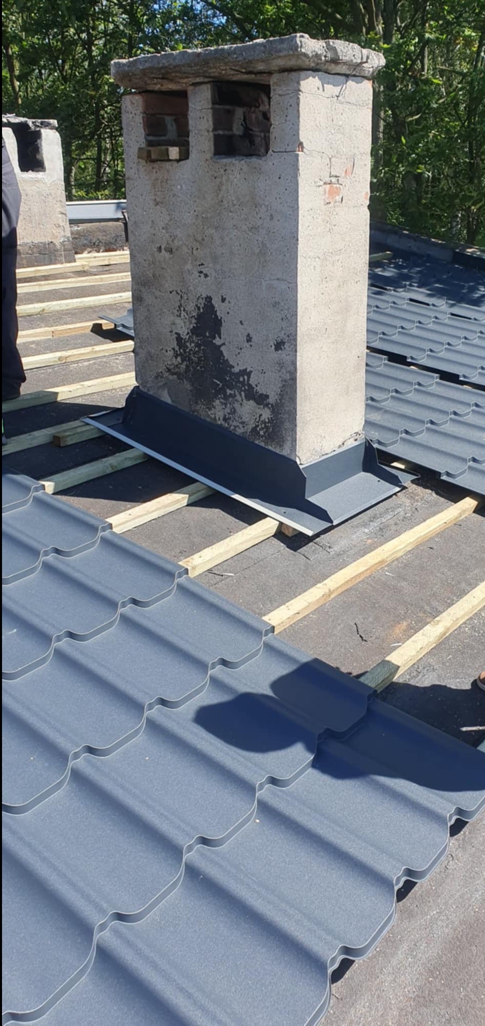 Montaż blaszanych paneli dachowych wokół betonowego komina na deskowaniu z drewnianych łat; widoczne ślady zabrudzeń na kominie.
