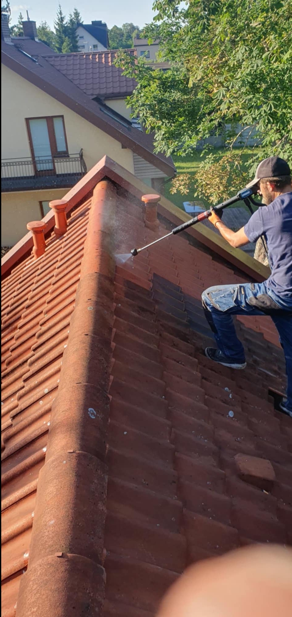 Mężczyzna w czapce czyści czerwoną dachówkę ceramiczną za pomocą myjki ciśnieniowej, widoczne zabrudzenia i strumień wody pod ciśnieniem, w tle drzewo i budynek mieszkalny.