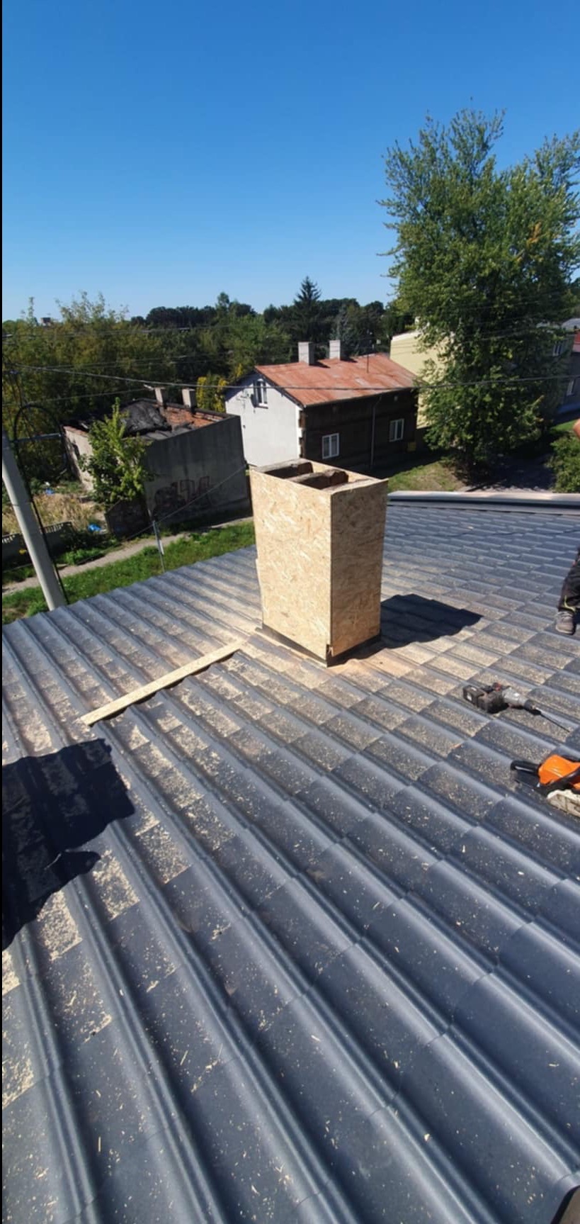 Szary dach z blachy trapezowej z drewnianą obudową komina w trakcie montażu, narzędzia dekarskie obok, widok na domy i zieleń w tle w słoneczny dzień.