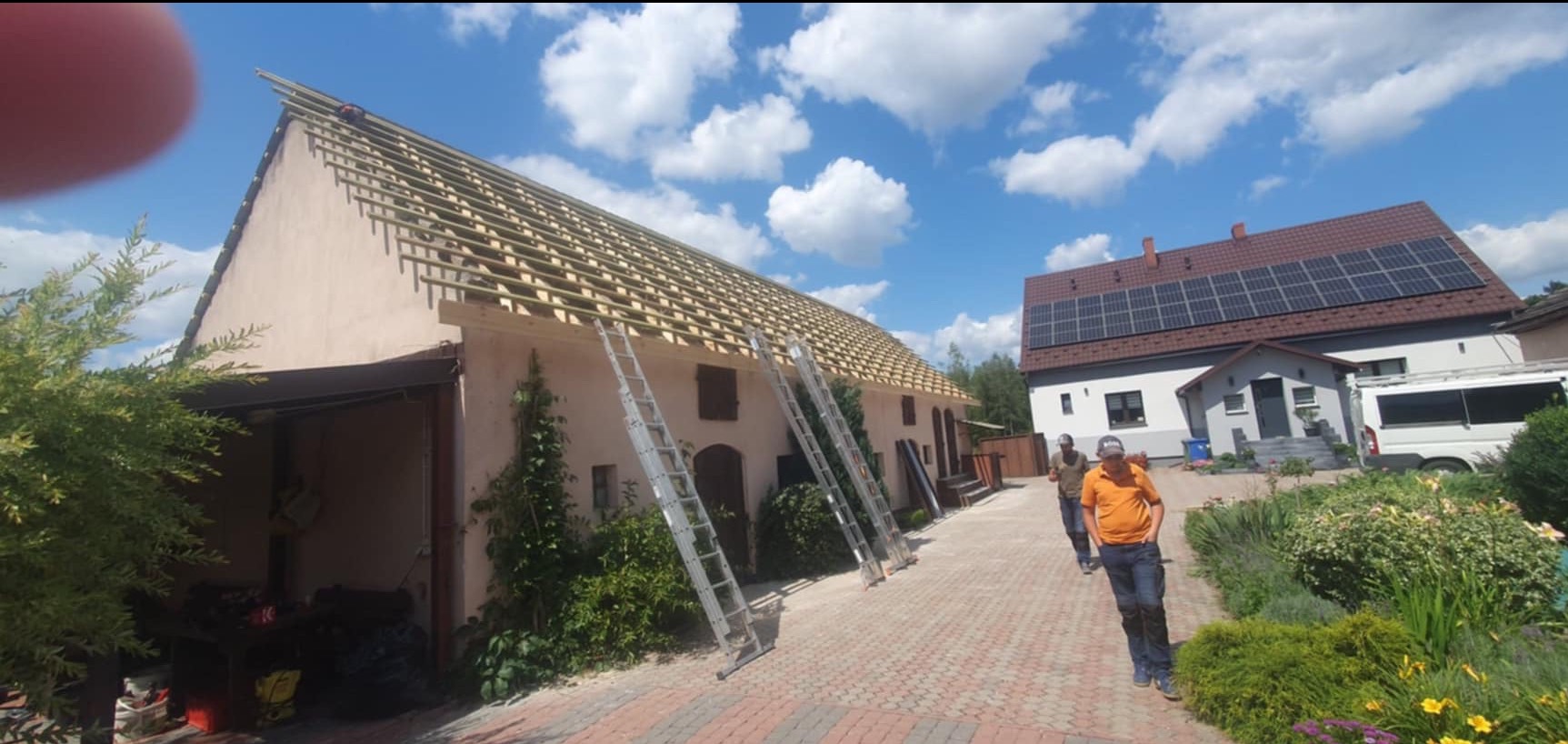 Widok z dołu na budynek z częściowo zdjętym poszyciem dachowym, widoczne krokwie i łaty, dwie przystawione drabiny, w tle drugi budynek z panelami słonecznymi na dachu, na pierwszym planie dwóch...