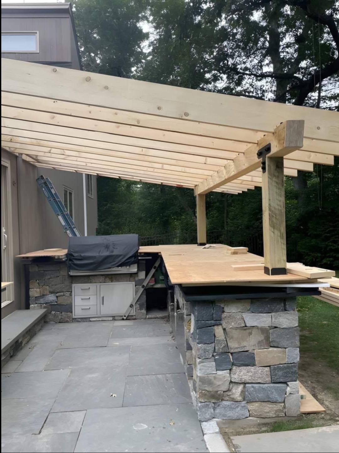 Wznoszenie drewnianej pergoli nad kamienną zabudową grilla na tarasie; widoczna drabina oparta o ścianę budynku.