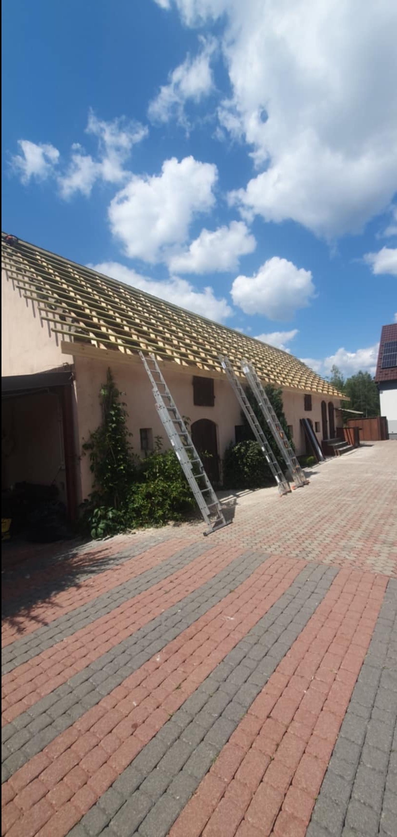 Budynek z odsłoniętą konstrukcją dachu, widoczne krokwie, dwie aluminiowe drabiny oparte o ścianę, kostka brukowa na podwórzu i błękitne niebo z chmurami w tle.