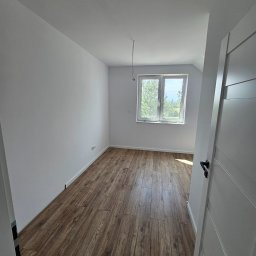 DB-BAU - Pusty pokój po remoncie z panelami podłogowymi w odcieniach brązu, świeżo pomalowane ściany na biało, białe okno z widokiem na zieleń i wystający kabel z sufitu.