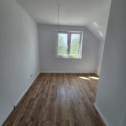 DB-BAU - Pusty pokój na poddaszu z drewnianą podłogą, białymi ścianami, oknem z widokiem na zieleń i wystającym przewodem z sufitu.