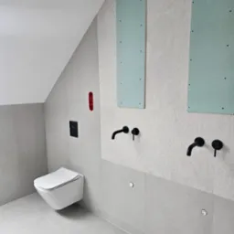 Wnętrze łazienki w Bolesławiu w trakcie remontu, z zamontowaną podtynkową toaletą, widoczne fragmenty płyt kartonowo-gipsowych na ścianie oraz czarne baterie umywalkowe.