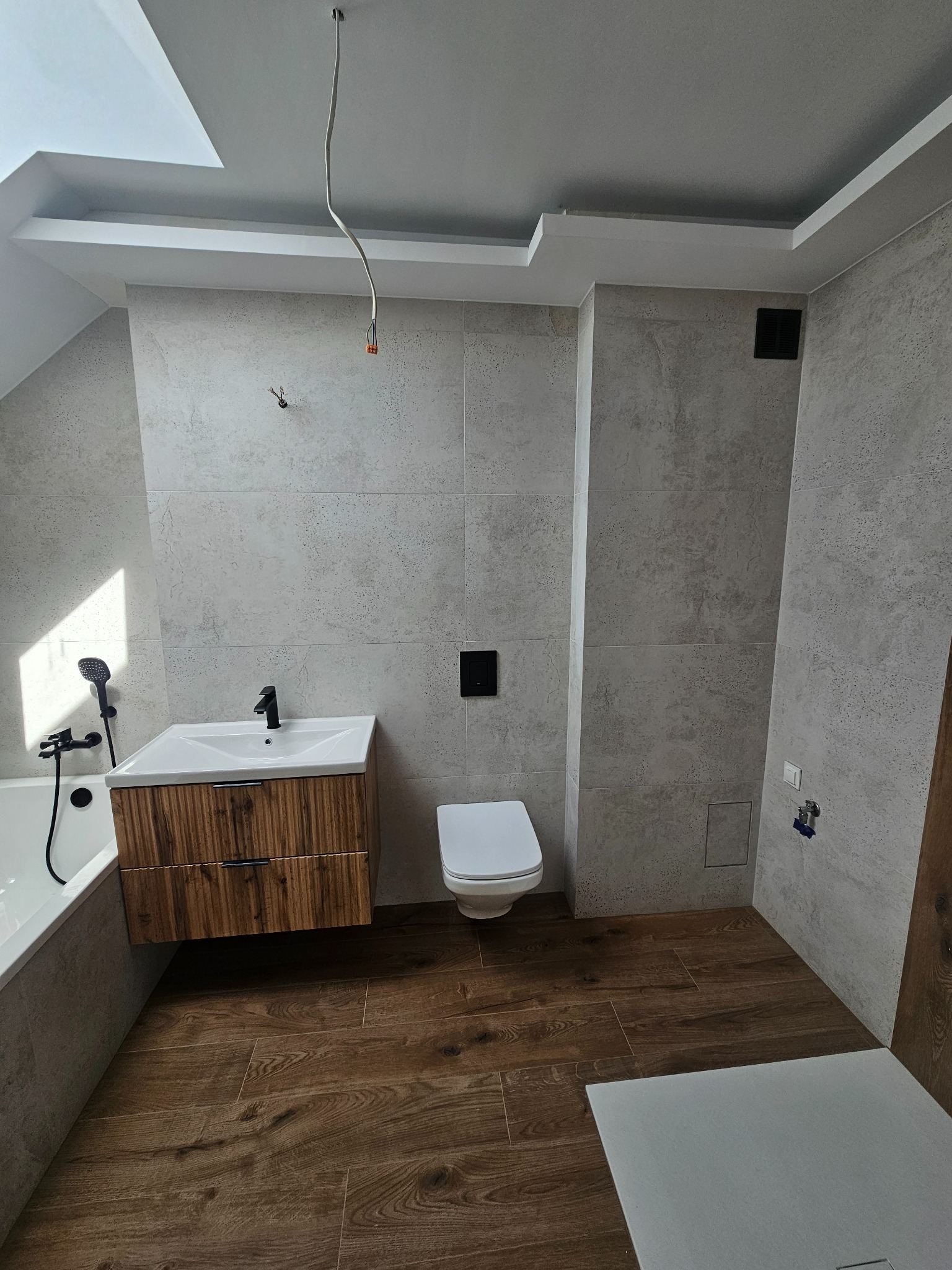Wykończona łazienka z szarymi płytkami imitującymi beton, drewnianą szafką pod umywalką, podwieszaną toaletą i drewnopodobną podłogą, widoczne niedokończone podłączenie elektryczne na suficie.